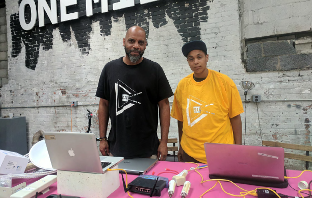 Digital stewards of DCTP Heru House and Gabrielle Knox. Photo via Jean Louis Farges. Image description: Two people stand behind a table covered with electronic equipment necessary to set up wireless internet.