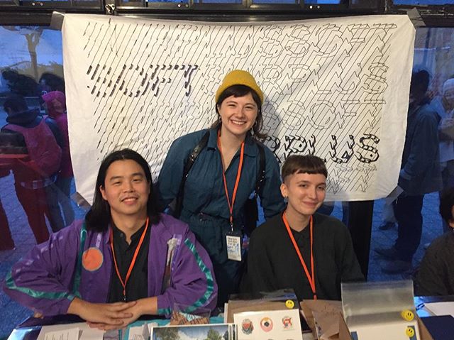 Soft Surplus members tabling at the New York Tech Zine Fair. Image description: Three people sit and stand behind a table filled with zines and other printed matters, happily smiling and looking at the camera.